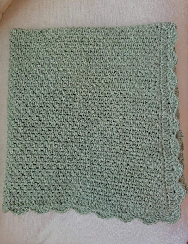 May include: A handmade, sage green crochet blanket. The blanket features a textured stitch pattern and a scalloped edge. The soft, muted green color and detailed craftsmanship suggest a cozy, comforting item.