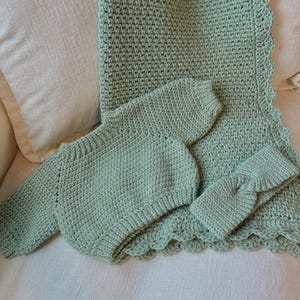 May include: A mint green crochet baby blanket, long-sleeved onesie, and headband set. The blanket has a scalloped edge. The set is displayed on a cream-colored surface. The crochet work has a textured appearance.