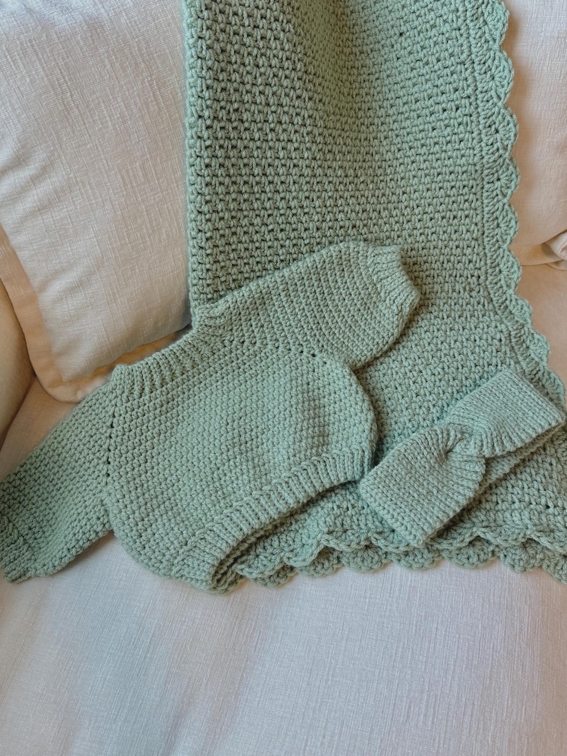 May include: A mint green crochet baby blanket, long-sleeved onesie, and headband set. The blanket has a scalloped edge. The set is displayed on a cream-colored surface. The crochet work has a textured appearance.