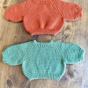 May include: Two handmade crochet sweaters in the image. One is a warm orange color, and the other is a soft green. Both sweaters have short sleeves and a round neckline. The sweaters are displayed on a wooden surface.