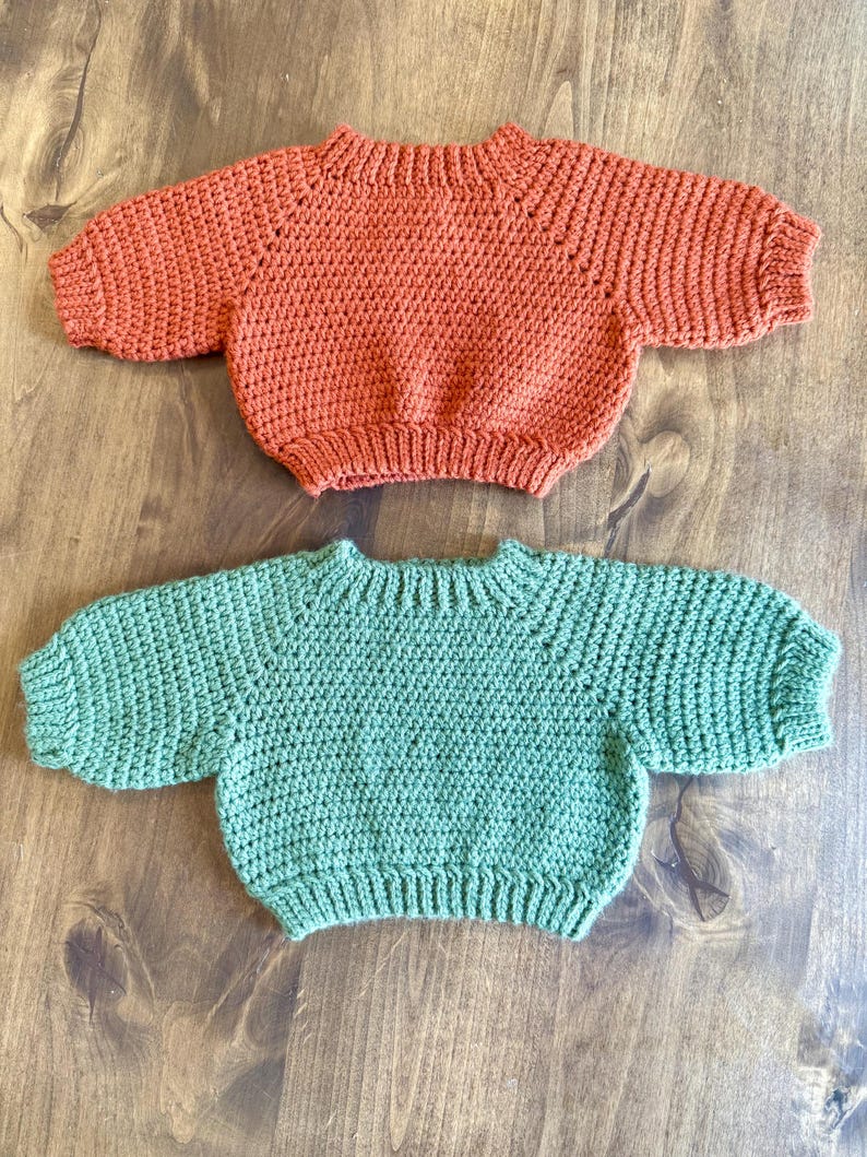 May include: Two handmade crochet sweaters in the image. One is a warm orange color, and the other is a soft green. Both sweaters have short sleeves and a round neckline. The sweaters are displayed on a wooden surface.