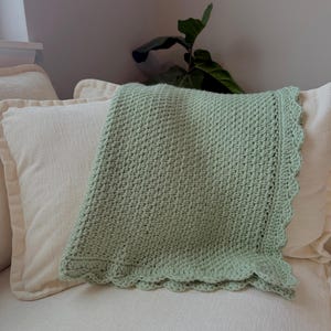 May include: A light green crocheted blanket with a scalloped edge is draped over two cream-colored pillows. The blanket has a textured pattern and is folded over. The pillows are on a cream-colored surface.