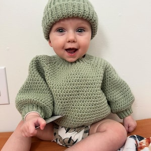 May include: A baby wearing a sage green crochet sweater and matching beanie. The baby is sitting on a brown surface and wearing a diaper with a leaf pattern. The baby is smiling and holding a small card.