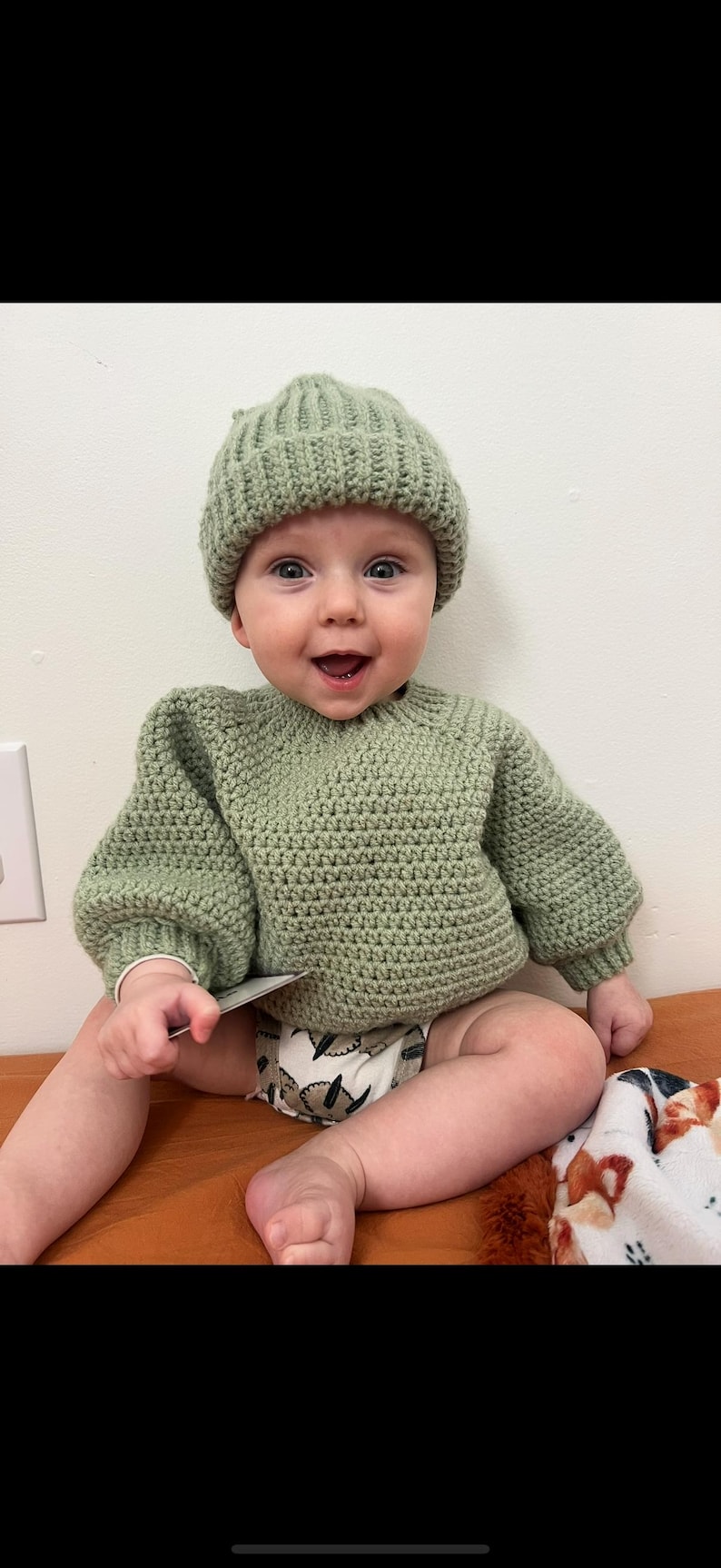 May include: A baby wearing a sage green crochet sweater and matching beanie. The baby is sitting on a brown surface and wearing a diaper with a leaf pattern. The baby is smiling and holding a small card.