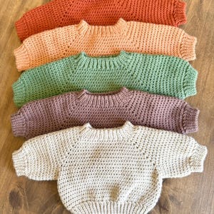 May include: Five handmade crochet sweaters in various colors: orange, peach, green, purple, and beige. Each sweater has a round neckline and long sleeves. The sweaters are arranged in a row on a wooden surface.