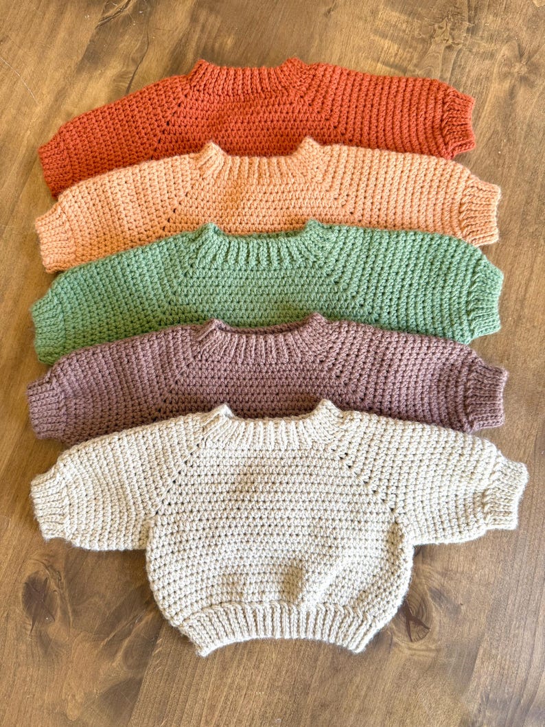 May include: Five handmade crochet sweaters in various colors: orange, peach, green, purple, and beige. Each sweater has a round neckline and long sleeves. The sweaters are arranged in a row on a wooden surface.