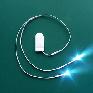 Könnte beinhalten: Eine weiße batteriebetriebene LED-Lichterkette mit zwei weißen Lichtern. Die Lichter sind eingeschaltet und strahlen ein helles weißes Licht ab.