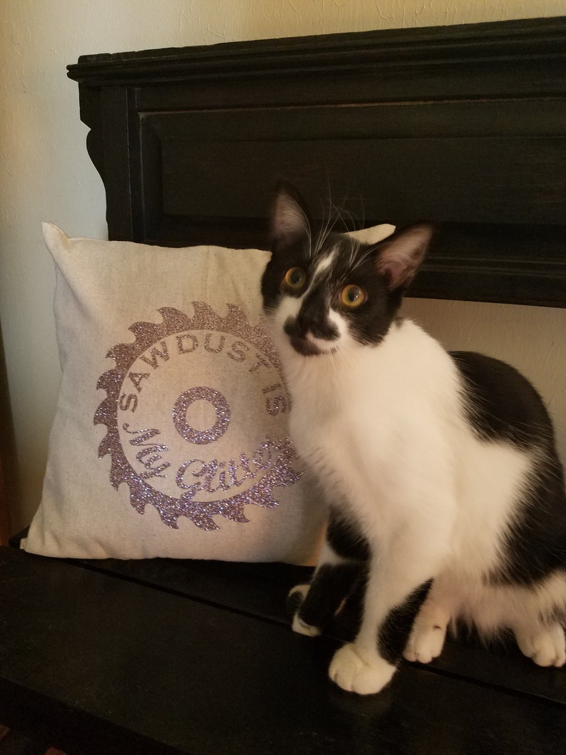 May include: A black and white cat sits on a bench with a white pillow. The pillow has a purple glitter design of a saw blade with the text "My Dust is Glitter".
