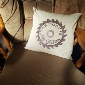 May include: A white throw pillow with a brown saw blade design that says "Sawdust is my glitter". The pillow is sitting on a brown armchair.