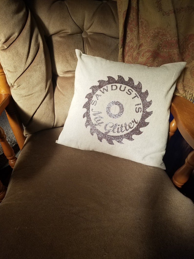 May include: A white throw pillow with a brown saw blade design that says "Sawdust is my glitter". The pillow is sitting on a brown armchair.
