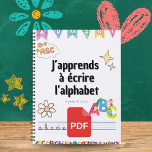 Può includere: Un quaderno a spirale con il titolo "J'apprends à écrire l'alphabet". La copertina presenta illustrazioni colorate, tra cui le lettere ABC, un fiore e un'icona PDF. Lo sfondo è una lavagna.