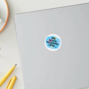May include: A round sticker with the text "ONE MENTAL BREAKDOWN LATER..." in blue and white on a light blue watercolor background. The sticker is on a grey laptop. A cup of coffee, gold binder clips, and a yellow pen are also visible.