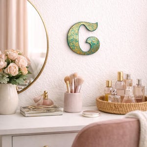 Puede incluir: Una letra decorativa "G" en color turquesa y dorado cuelga sobre un tocador. El tocador presenta un espejo redondo, flores rosas, brochas de maquillaje, frascos de perfume y una silla rosa. El tema general es la belleza y la decoración del hogar.
