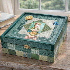 May include: A decorative wooden box with a green and white patchwork design. The lid features a sun, a portrait, and script. The box is on a wooden surface, with a book and seashells nearby.