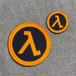 May include: Two circular patches featuring a stylized orange lambda symbol on a navy blue background, with an orange border. One patch is larger than the other. The patches are likely for clothing or accessories.
