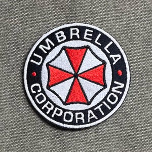 Może przedstawiać: Okrągła haftowana naszywka z logo Umbrella Corporation. Wzór zawiera centralną ikonę czerwono-białego segmentowanego parasola, otoczoną czarną obwódką z białym napisem UMBRELLA CORPORATION oraz dwiema małymi czerwonymi kropkami.