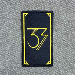 Könnte beinhalten: Ein rechteckiger, schwarz bestickter Aufnäher mit gelbem Rand und der Zahl 33 in einer stilisierten gelben Schriftart. Das Design umfasst dünne gelbe Eckakzente und eine Doppellinien-Ästhetik für die Zahl und den Rahmen.