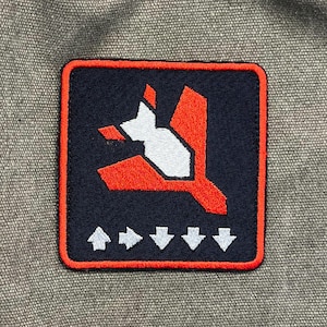 May include: Embroidered patch with a navy blue background and a red border. The patch features a white and red graphic design of a stylized airplane and arrows. The patch is square and has a textured appearance.