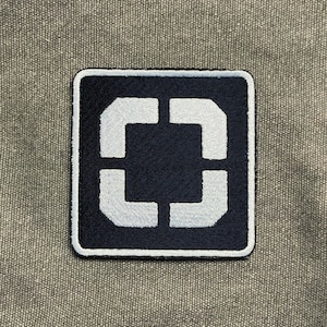 May include: Square patch with a navy blue background and a white, stylized, open square design. The patch has a white border and is set against a textured, gray-green fabric.