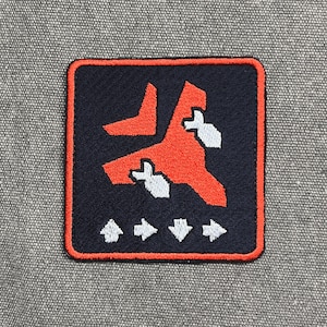 May include: Embroidered patch with a navy blue background and a red and white graphic design. The patch has a red border and four white arrows at the bottom. The patch is square and is set against a gray fabric background.