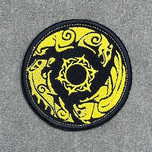 Op de afbeelding: Een ronde geborduurde patch met een zwarte achtergrond en een felgeel ontwerp. Het ontwerp bestaat uit drie gestileerde dierenfiguren die in een cirkelvormig patroon zijn gerangschikt rond een centraal zonachtig embleem met puntige stralen.