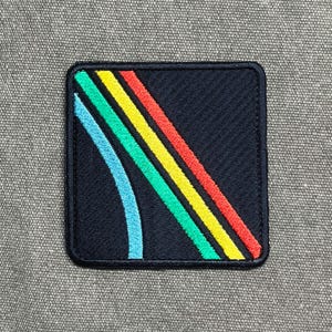 May include: Square patch with a black background and a colorful design. The design features a curved blue line and three diagonal stripes in green, yellow, and red. The patch has a black border.