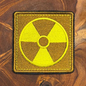Könnte beinhalten: Quadratischer Aufnäher mit goldenem Rand und einem Strahlungssymbol in der Mitte. Das Strahlungssymbol ist gelb und hat drei gelbe Dreiecke, die von einem zentralen gelben Kreis ausgehen. Der Hintergrund ist ein dunkleres Gold.