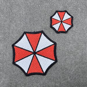 Op de afbeelding: Twee geborduurde patches met een achthoekig parapluontwerp met afwisselende rode en witte driehoekige segmenten. Elke patch is omlijnd met een dikke zwarte rand, wat een geometrisch patroon creëert met acht secties die vanuit het midden uitstralen.