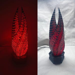 Peut inclure: Une lampe sculpturale haute à deux pointes avec un extérieur sombre et texturé composé de segments horizontaux. La structure est éclairée de l'intérieur, projetant une lueur rouge vive à travers des symboles extraterrestres gravés.