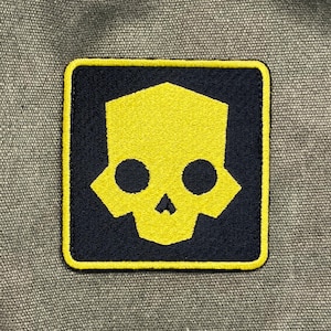 May include: Square patch featuring a stylized yellow skull design on a navy blue background. The skull has two circular eye sockets and a triangular nose. The patch is bordered with a yellow trim and is set against a gray fabric.