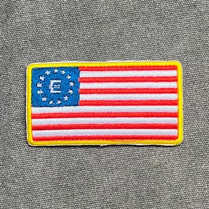 May include: Rectangular patch with a stylized American flag design. The flag has red and white stripes, a blue canton with white stars, and a yellow border. The letter "E" is centered in the blue canton. The patch is likely for clothing or accessories.