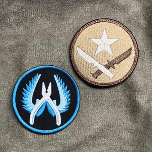 Könnte beinhalten: Zwei runde, gestickte Aufnäher. Einer hat einen schwarzen Hintergrund mit einem blau-weißen Design. Der andere hat einen beigefarbenen Hintergrund mit einem weißen Stern und gekreuzten Messern. Beide Aufnäher haben einen gestickten Rand.