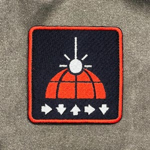 May include: Embroidered patch with a navy blue background and a red and white graphic. The graphic features a stylized globe, arrows, and a sunburst design. The patch has a red border and is on a gray fabric surface.