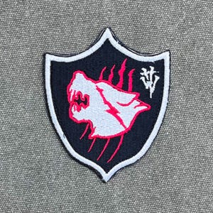Op de afbeelding: Een schildvormige geborduurde patch met een zwarte achtergrond en een witte rand. Het ontwerp toont een wit silhouet van een wolvenkop omlijnd in felroze, geaccentueerd door roze klauwsporen en een wit gestileerd symbool in de rechterbovenhoek.