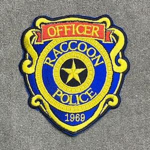 Puede incluir: Parche bordado con un diseño de insignia de policía azul y amarillo. El parche dice "OFFICER RACCOON POLICE 1969" en letras amarillas y blancas. Una estrella dorada está en el centro de la insignia.