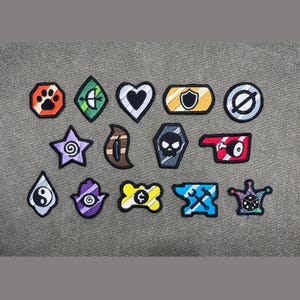 Op de afbeelding: Een verzameling van veertien geborduurde patches met diverse vormen en symbolen, waaronder een pootafdruk, hart, schedel, aambeeld en dobbelstenen. Elke patch heeft een zwarte rand en kleurrijk borduurwerk met iconen zoals een boog, schild, yin-yang en hamsa-hand.