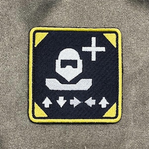 May include: A square patch with a black background and a yellow border. The patch features a white graphic of a person with a plus sign and arrows. The patch is likely an emblem or a badge.