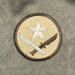 Könnte beinhalten: Runder, gestickter Aufnäher mit einem weißen Stern und zwei gekreuzten Messern auf beigem Hintergrund. Der Aufnäher hat einen braunen Rand. Der Aufnäher befindet sich auf einem strukturierten, olivgrünen Stoff.