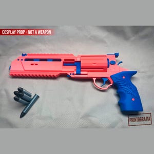 Könnte beinhalten: Ein rosafarbener und blauer Cosplay-Revolver mit strukturiertem Griff, Flügeldetails und einer oberen Schiene. Das Set enthält vier zubehörteile in Patronenform mit schwarzen Spitzen. Der Text COSPLAY PROP - NOT A WEAPON ist sichtbar.