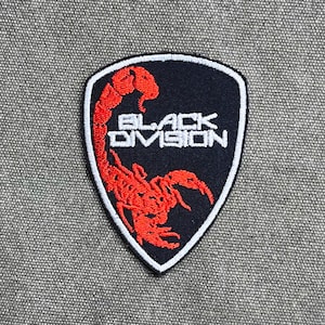 Op de afbeelding: Een schildvormige geborduurde patch met een zwarte achtergrond en een witte rand. De patch bevat een rode schorpioengrafiek en de witte tekst "BLACK DIVISION" in het midden van het ontwerp, met de staart van de schorpioen omhoog gebogen.