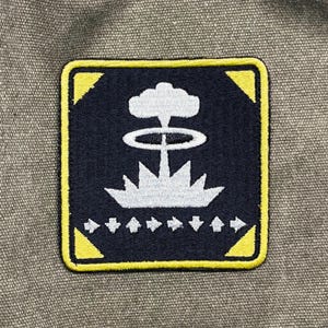 May include: Square patch with a navy blue background and a yellow border. The patch features a white graphic of an atomic explosion. Below the explosion are white arrows pointing to the right.