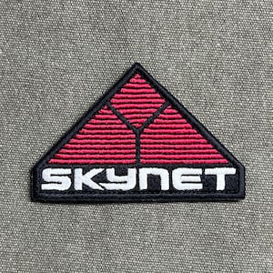 May include: A triangular embroidered patch featuring a black border and a red upper section with horizontal lines forming a Y-shape. The lower section is black with the word SKYNET stitched in bold white capital letters across the bottom.