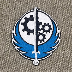 May include: Embroidered patch with a white circle, black gears, and a sword design, divided by a black sword. The design is set against a blue wing-like shape with a white border. The patch is on a textured grey background.