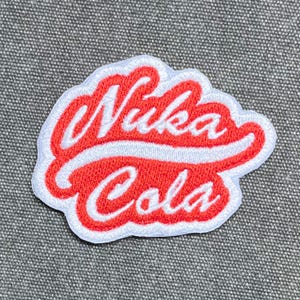 Puede incluir: Un parche bordado que presenta el texto Nuka Cola en una fuente cursiva blanca. Las letras están sobre un fondo rojo brillante con un borde blanco, creando un diseño de logotipo distintivo que sigue los contornos del texto en blanco.
