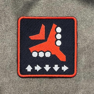 May include: Embroidered patch with a navy blue background and a red design. The design includes a stylized arrow, three white circles, and four arrows. The patch has a red border and is set against a gray fabric.