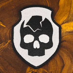 May include: Embroidered patch with a black skull design and a cracked top, set against a white shield-shaped background. The patch has a detailed, stitched appearance, suitable for application on clothing or accessories.