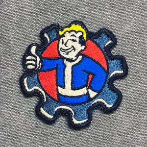 Op de afbeelding: Geborduurde patch met een lachend stripfiguur dat een duim omhoog geeft. Het personage draagt een blauwe overall met een witte kraag, tegen een rode cirkel. De patch heeft de vorm van een blauw tandwiel met een zwarte rand.
