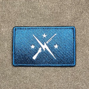 Op de afbeelding: Een rechthoekige blauwe patch met een wit ontwerp met een bliksemschicht, gekruiste lijnen en drie sterren. De patch heeft een gestikte rand en is geplaatst tegen een getextureerde grijze achtergrond.