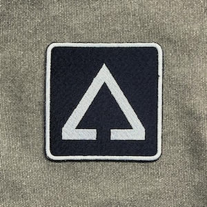 May include: A square patch with a navy blue background and a white arrow-like design. The patch has a white border and is set against a textured, gray-green fabric. The design is simple and geometric.