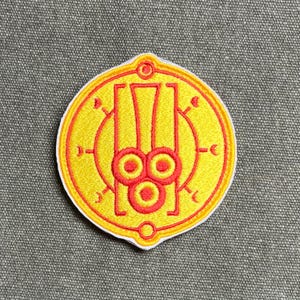 Peut inclure: Un écusson brodé circulaire présentant un fond jaune vif avec un motif géométrique rouge. Le centre affiche une forme rectangulaire verticale avec trois cercles rouges à la base, entourée d'une fine bordure circulaire rouge avec des détails.
